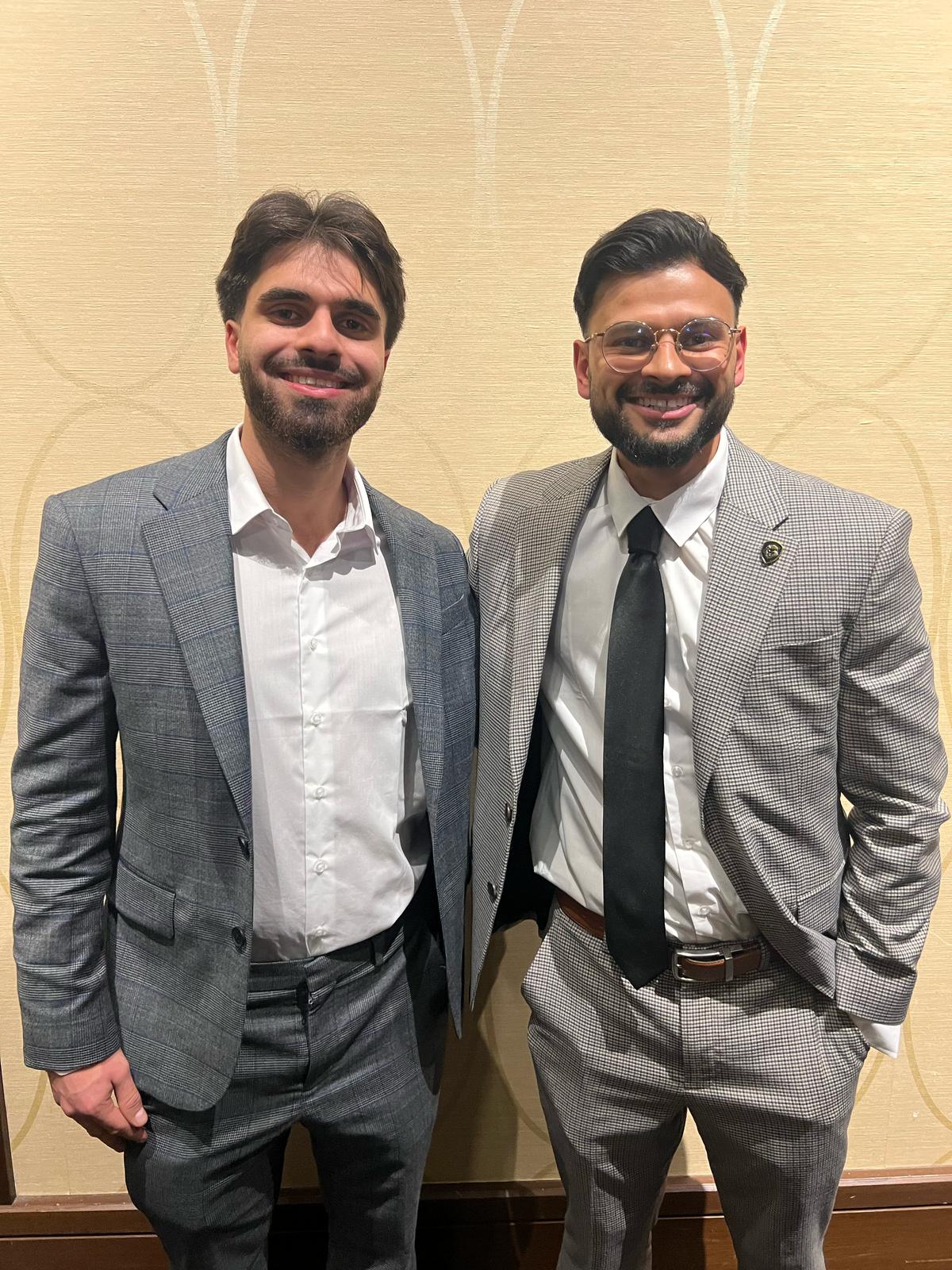 Two young businessmen in tailored grey and houndstooth suits smiling for a professional portrait at a marketing industry event.